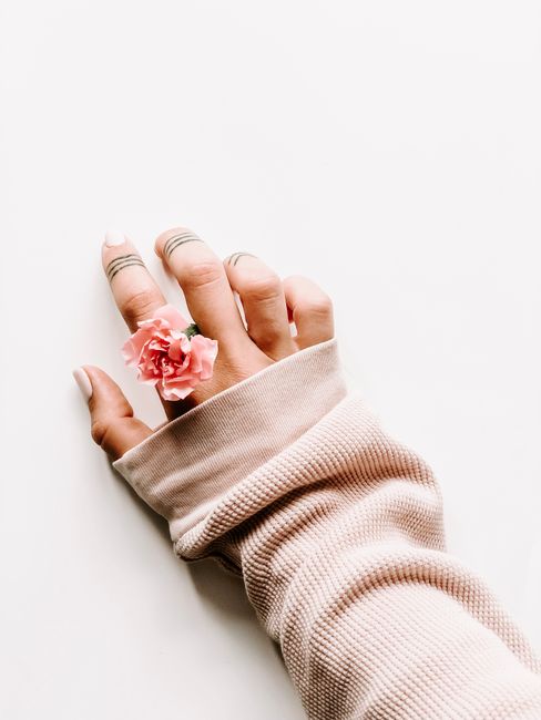 A delicate hand wearing a pink flower ring and cozy sweater against a white backdrop, showcasing minimalist style.