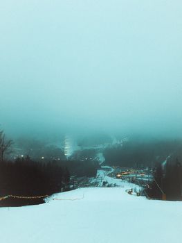 A snowy ski slope descends into a foggy valley with lit up village in winter twilight.