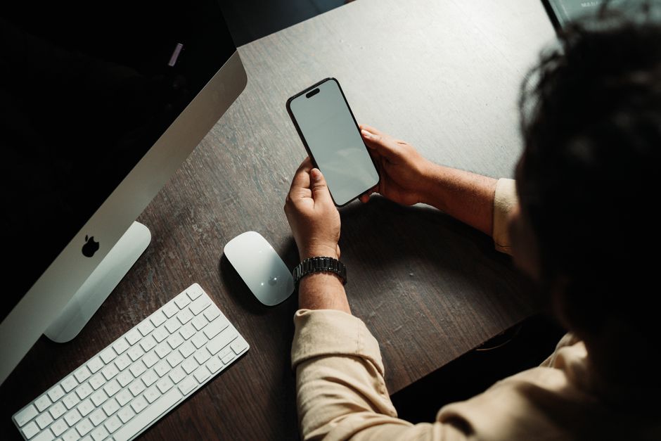Photo by Aathif Aarifeen on Pexels A person looking at a smartphone screen while sitting at a desk with computer and mouse.