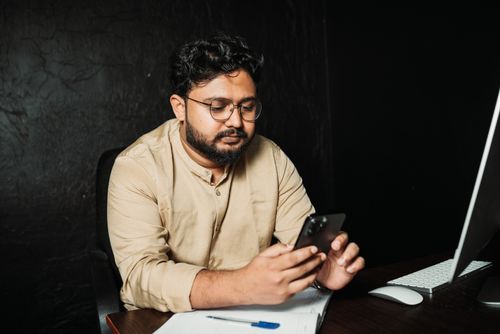 Free Man with beard using a smartphone at an office desk, focused and working. Stock Photo