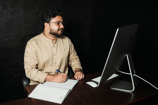 Man in an office setting writing in notebook while working on a desktop computer.