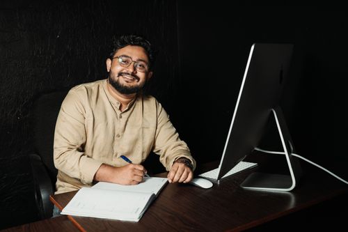 Free Business professional smiling at desk with computer and notebook, portraying a modern office setup. Stock Photo