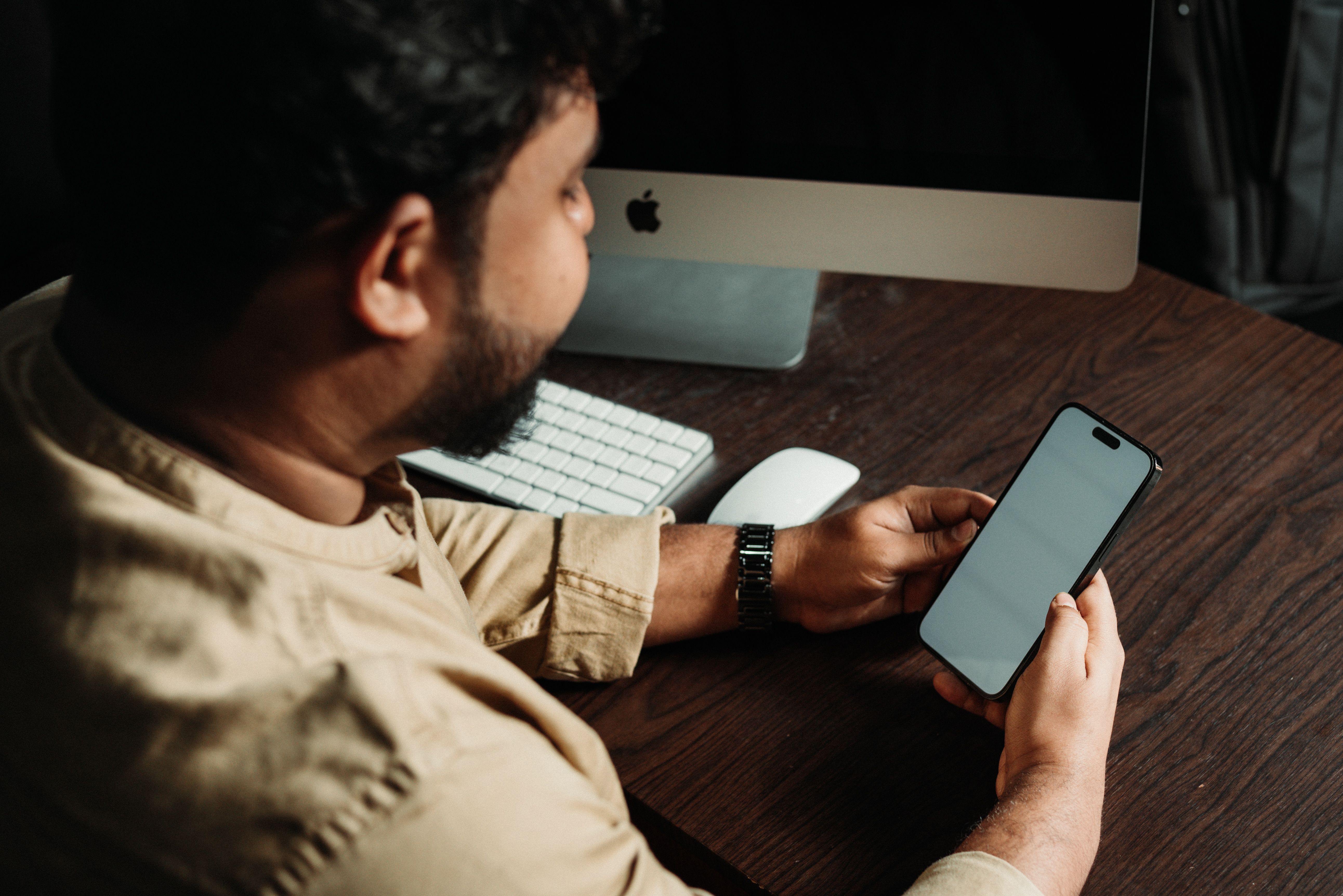 Adult male browsing smartphone at office desk with computer setup.