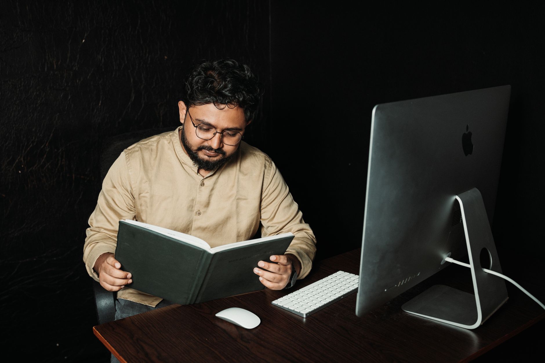 A man reading a book while seated at a desk with a computer, showcasing a blend of technology and traditional learning.