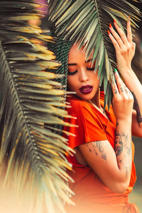 бесплатная Женщина в оранжевой рубашке стоит под листьями кокосовых пальм Стоковое фото