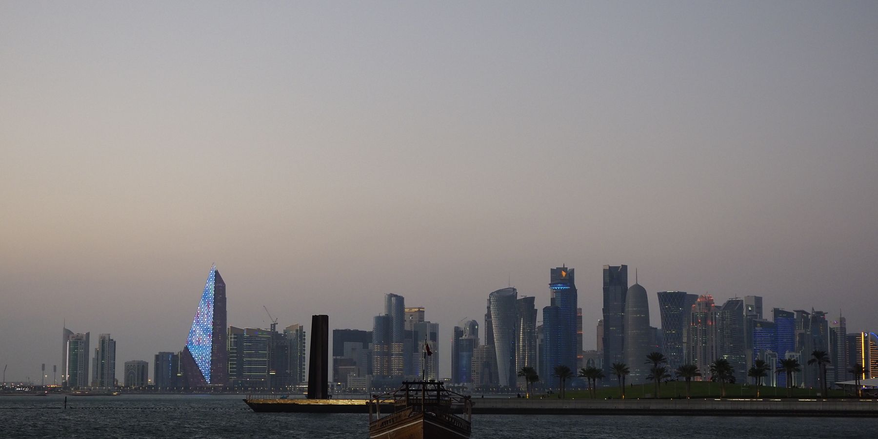 Scenic view of Doha city skyline with traditional dhow boat at dusk along the waterfront.