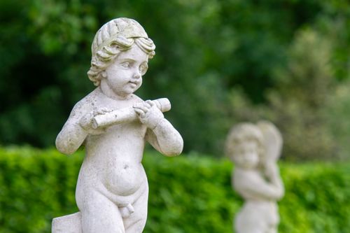 Free Elegant marble cherub sculptures set against a lush, green garden background in summer. Stock Photo