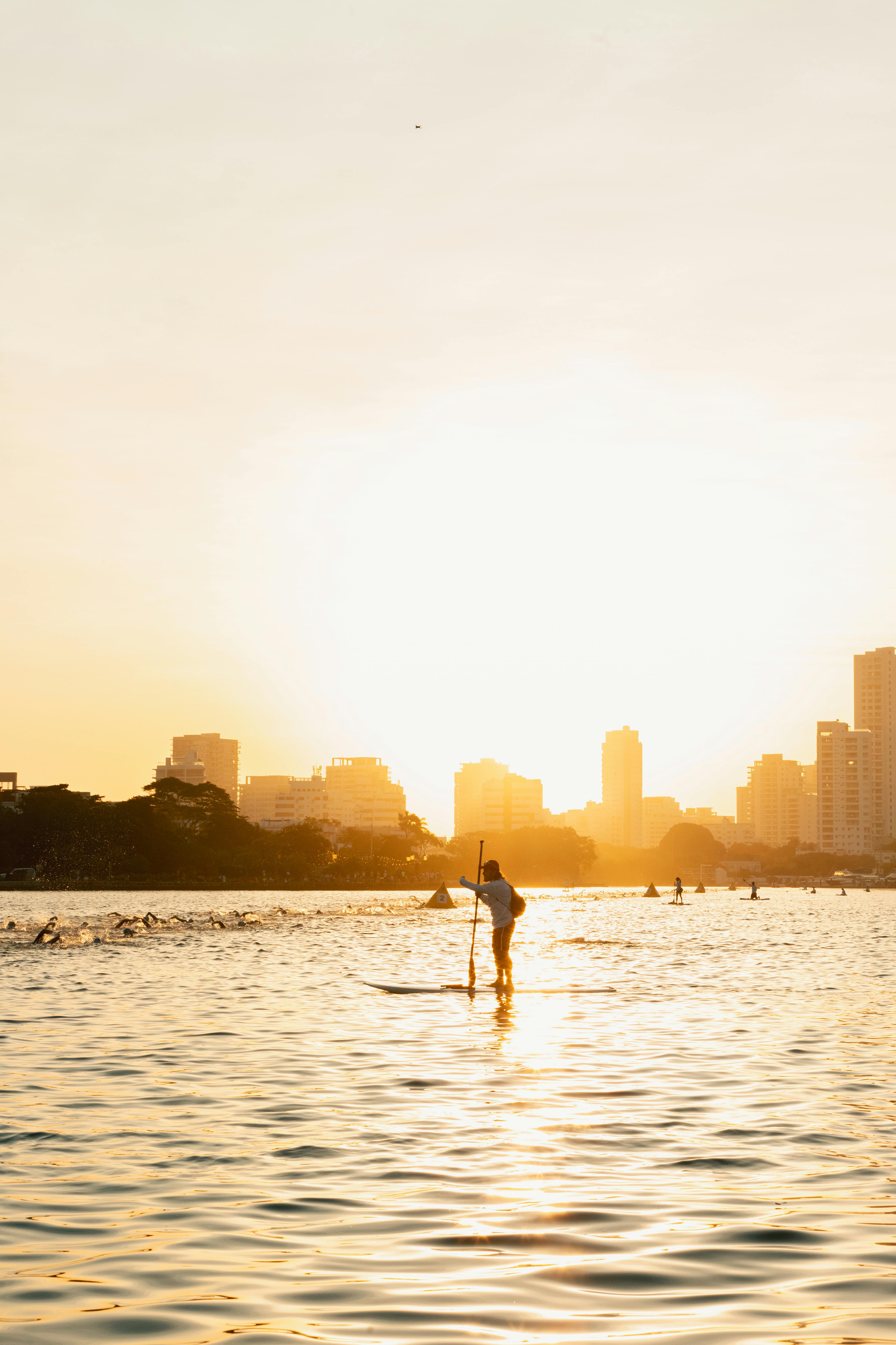 A man paddleboards on a calm urban lake with a city skyline at sunset, creating a serene silhouette.