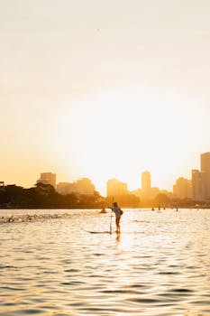 A man paddleboards on a calm urban lake with a city skyline at sunset, creating a serene silhouette.