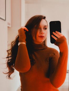 Woman in a brown knit sweater taking a mirror selfie indoors with a smartphone.