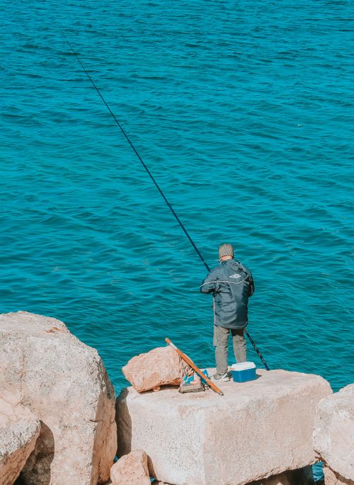 Free A man stands on boulders fishing with a rod along the Alexandria seashore in Egypt. Stock Photo