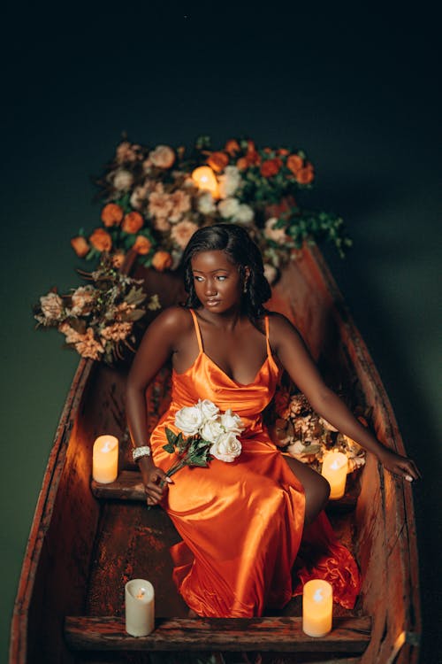 Young Woman in an Orange Evening Dress with a Slit on a Wooden Boat Among Flowers and Lit Candles