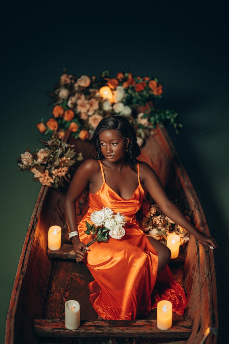 Young Woman In An Orange Evening Dress With A Slit On A Wooden Boat Among Flowers And Lit Candles