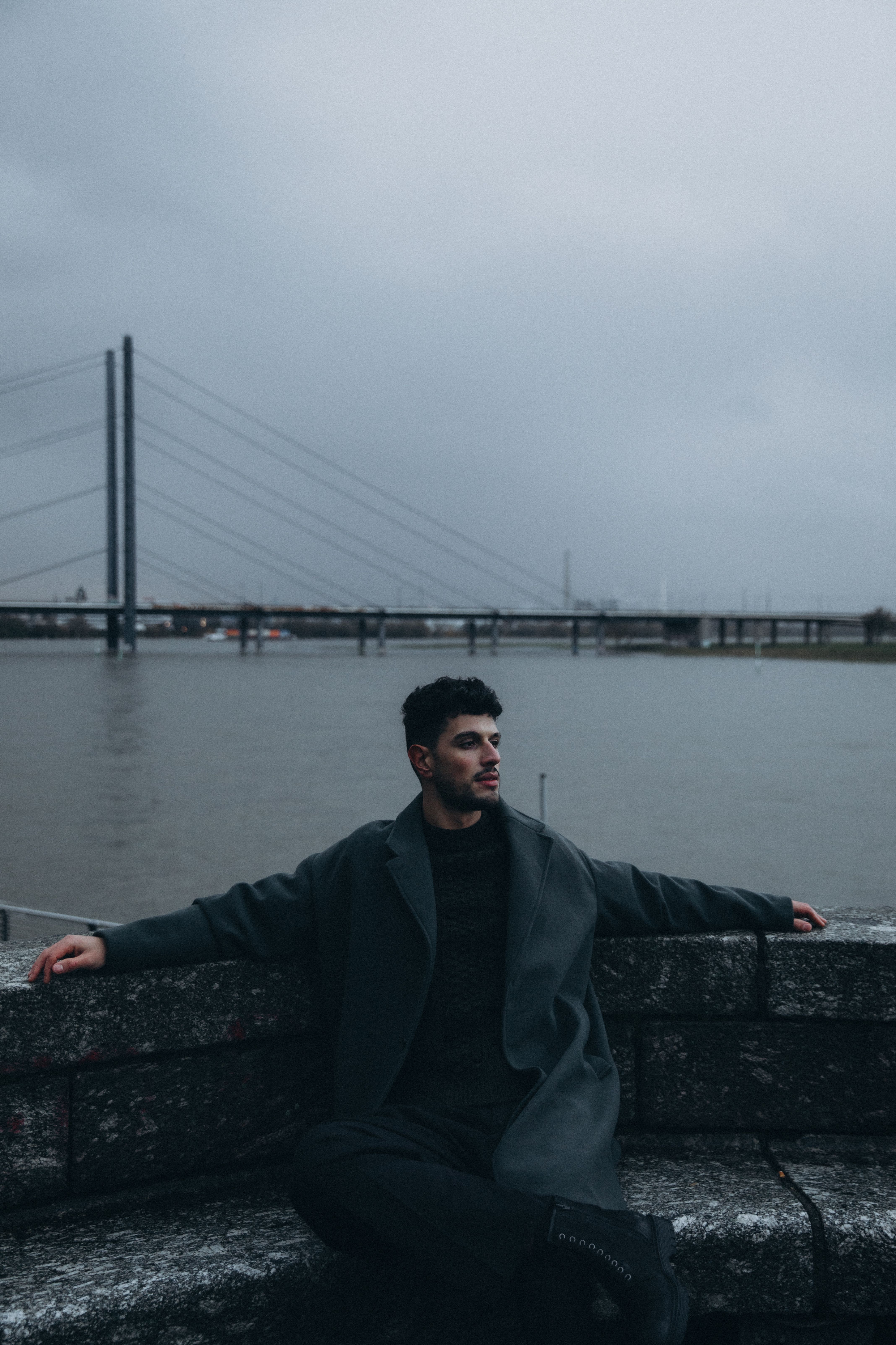 A man in a coat sits calmly by the waterfront, overlooking a city bridge under overcast skies.