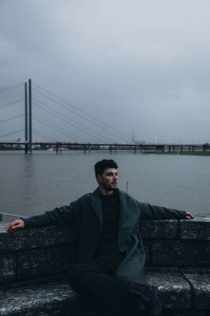 A man in a coat sits calmly by the waterfront, overlooking a city bridge under overcast skies.