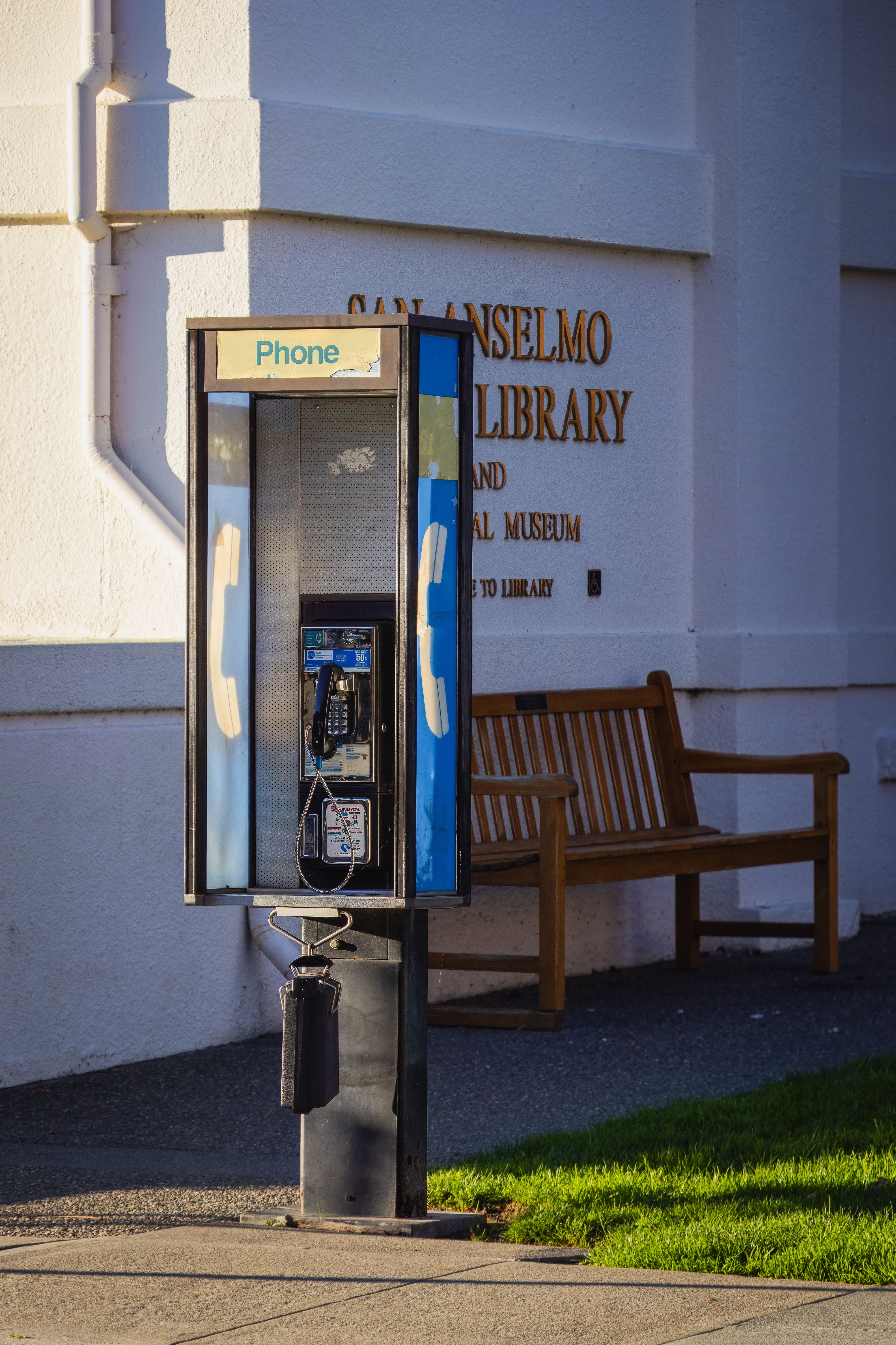 Classic payphone booth outside San Anselmo Library with a wooden bench nearby.