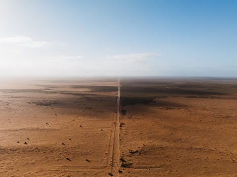 Aerial shot showcasing a long dirt road cutting through a vast desert landscape under a bright blue sky.