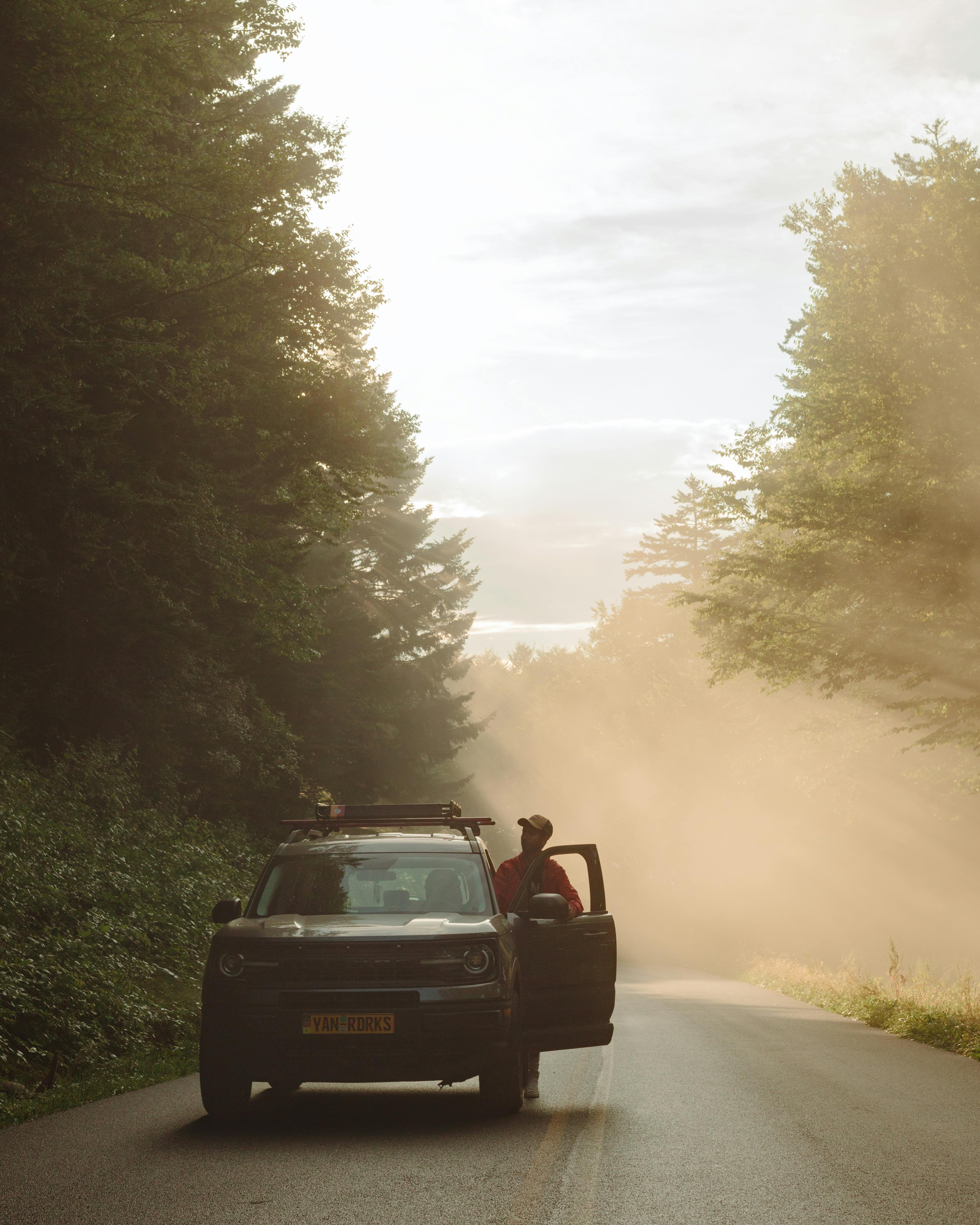 A car on a foggy forest road with a person standing by the door amidst the morning sunlight.