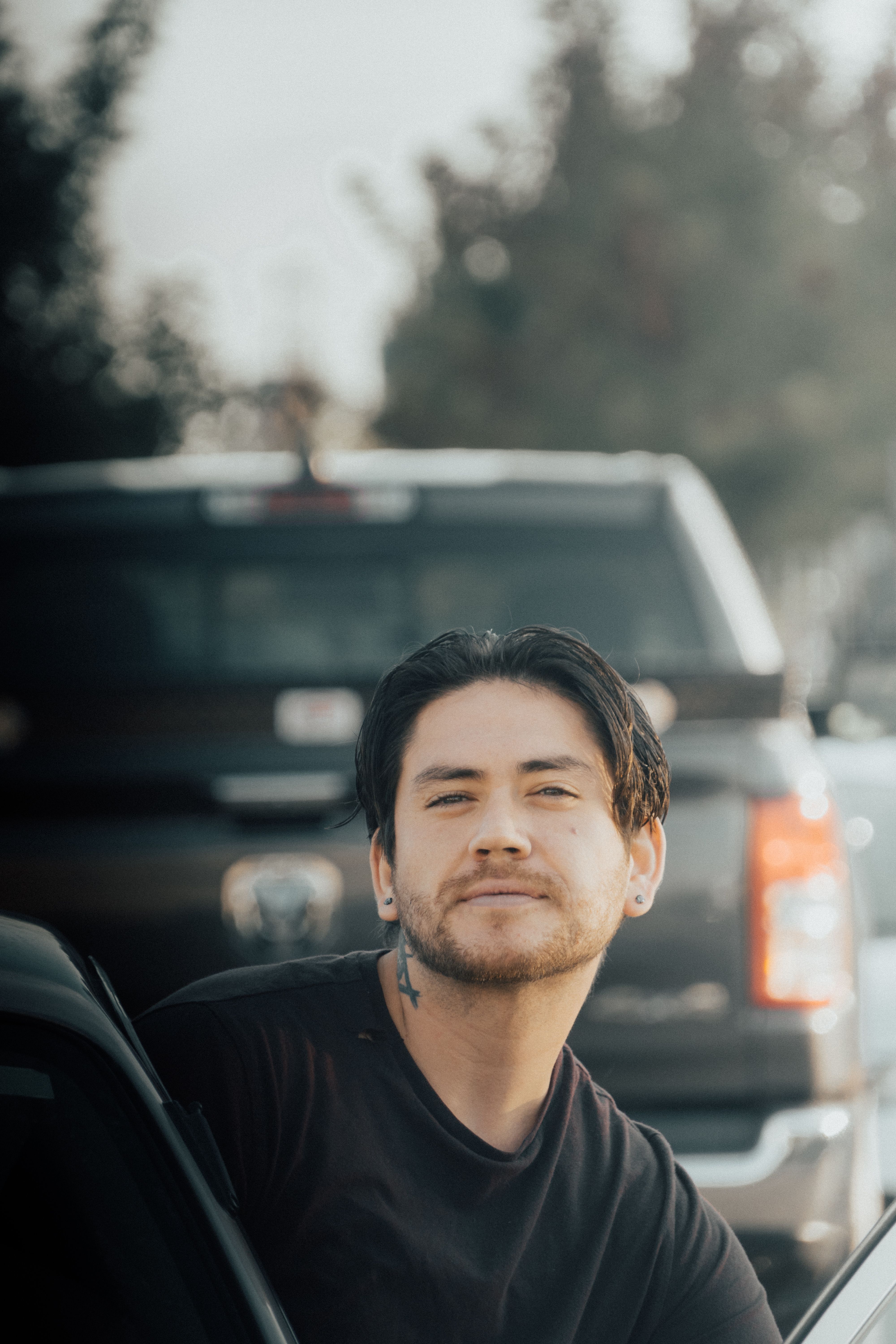 A young tattooed man gazes into the camera while standing near a car outdoors.