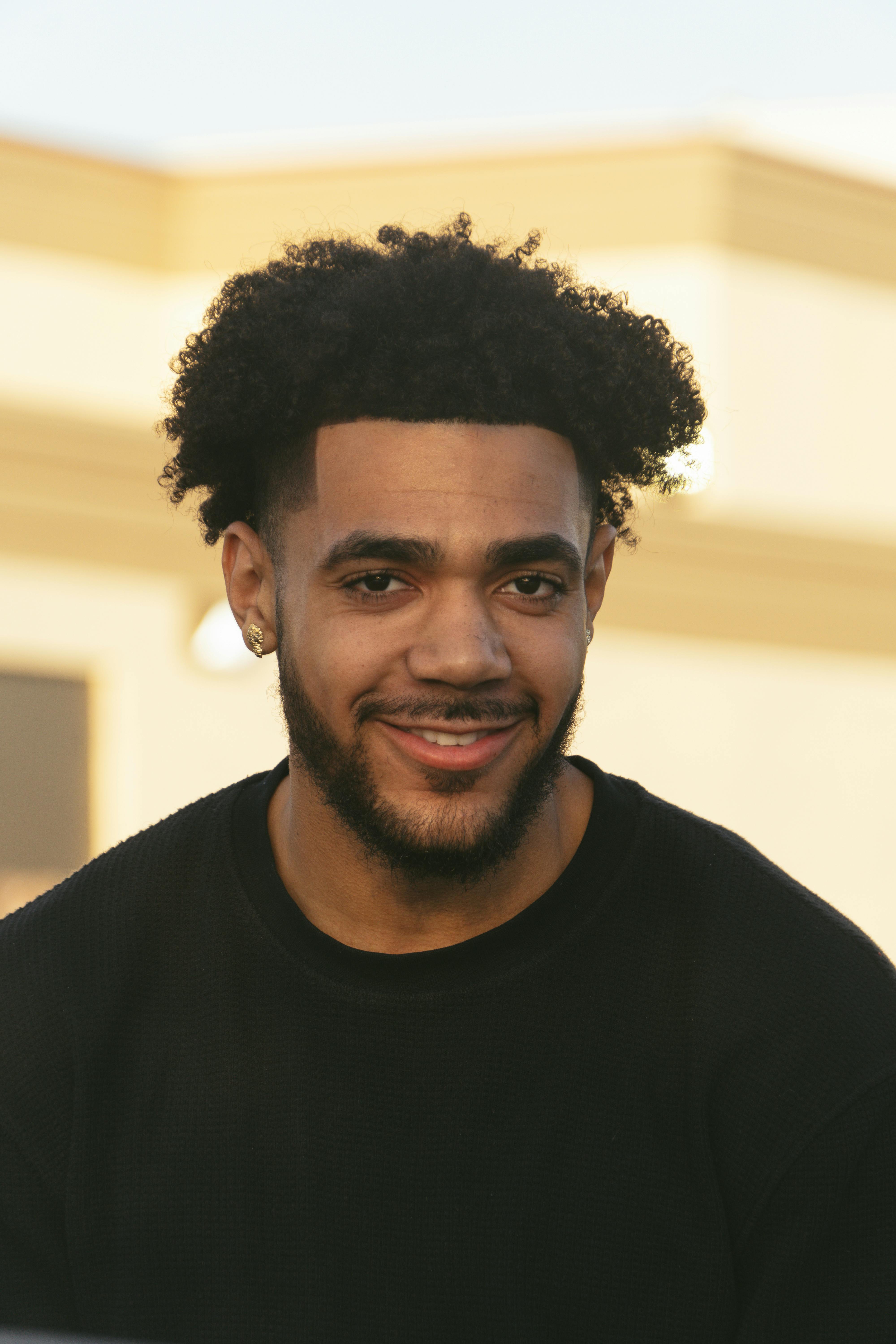 Portrait of a smiling man with curly hair and beard outdoors, showcasing casual fashion.