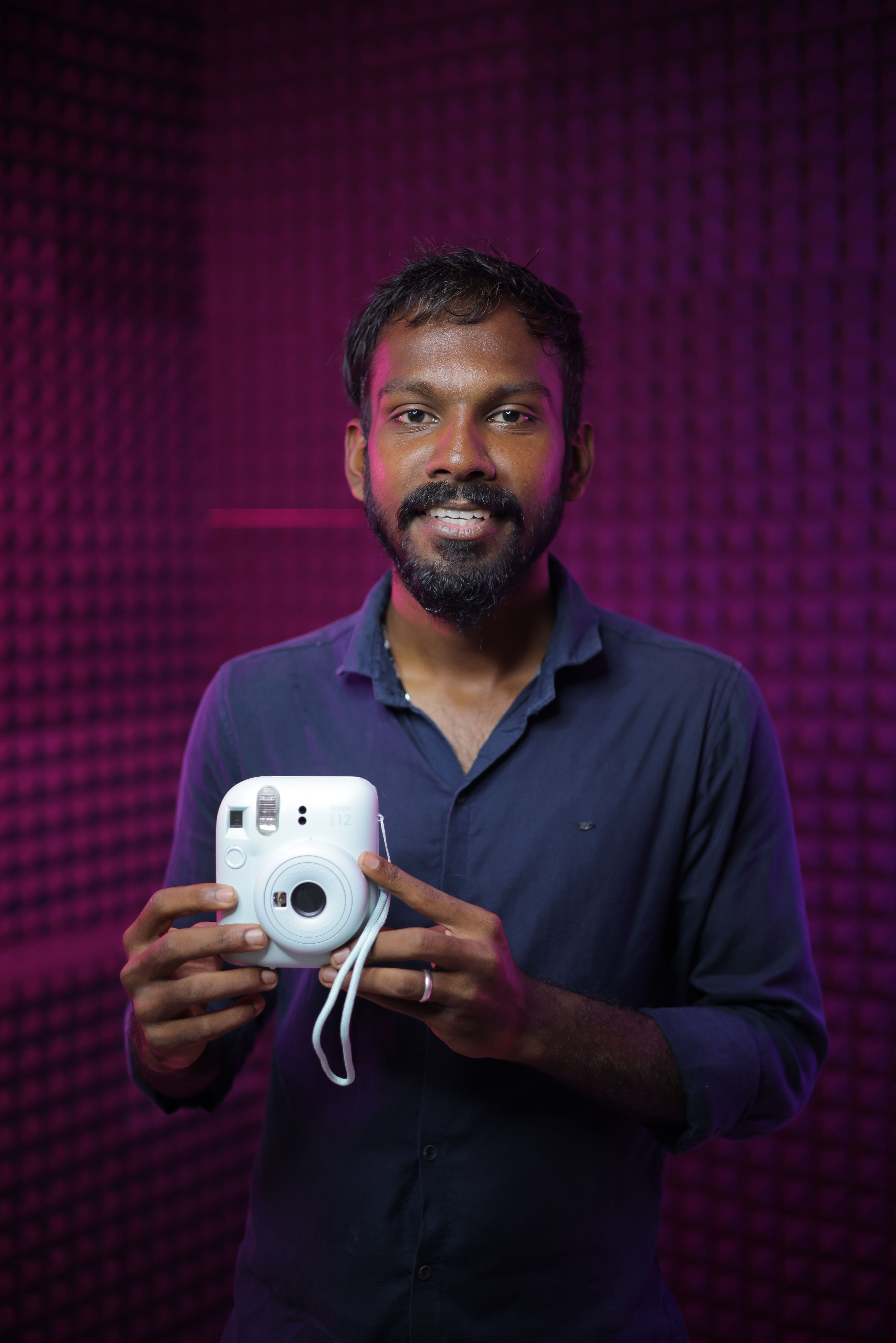 A smiling man holding a white camera with a vibrant purple background.