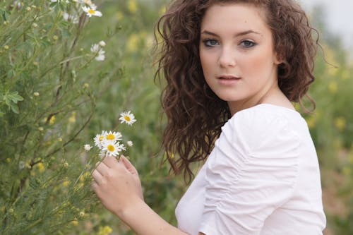 Young Woman in a White Blouse Standing on a Meadow and Touching a Flower
