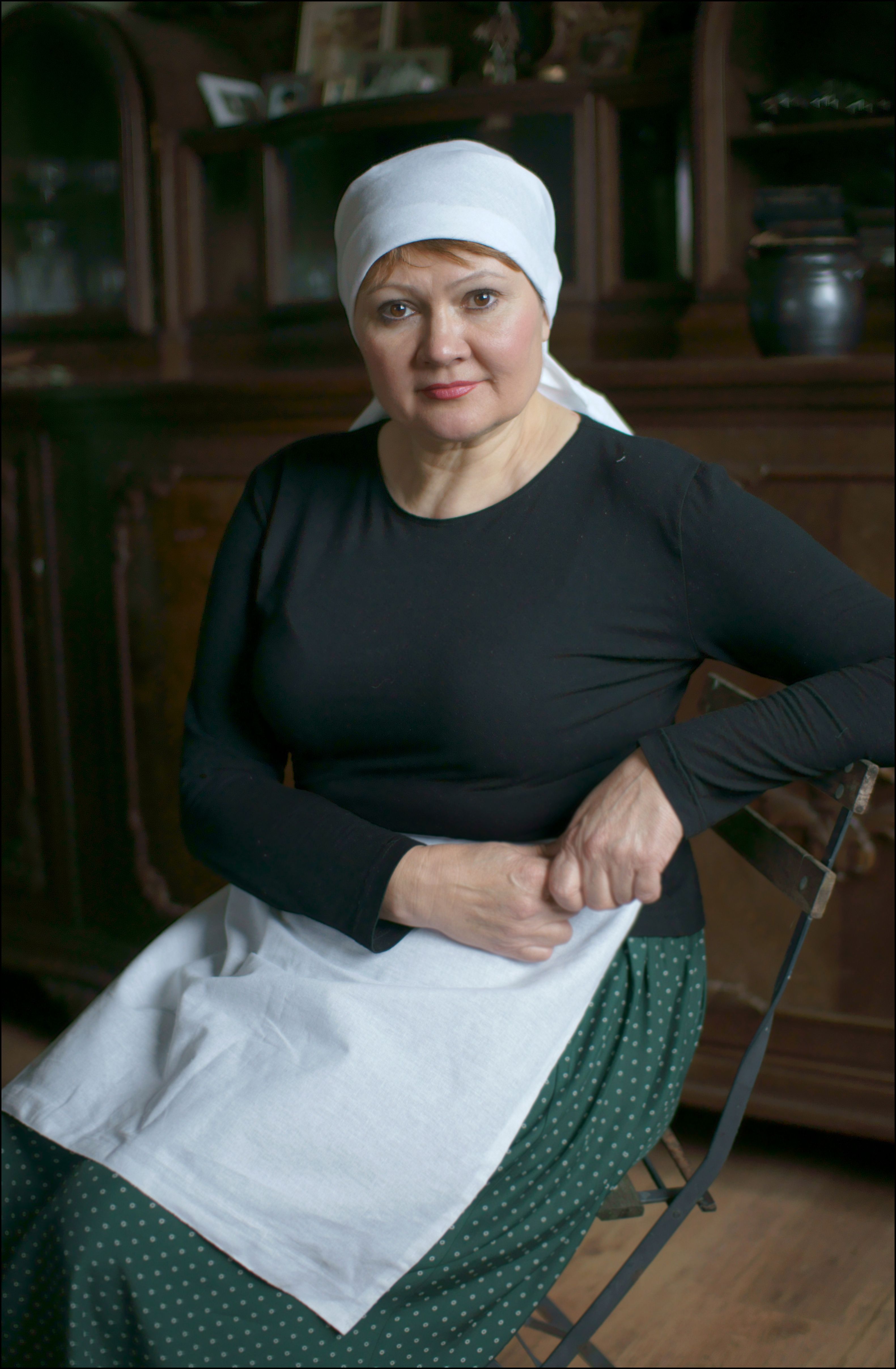 Elegant portrait of a senior woman wearing a headscarf and apron sitting indoors.