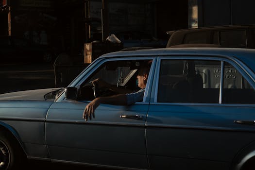 Man driving a classic car through city streets during sunset, showcasing retro urban life.