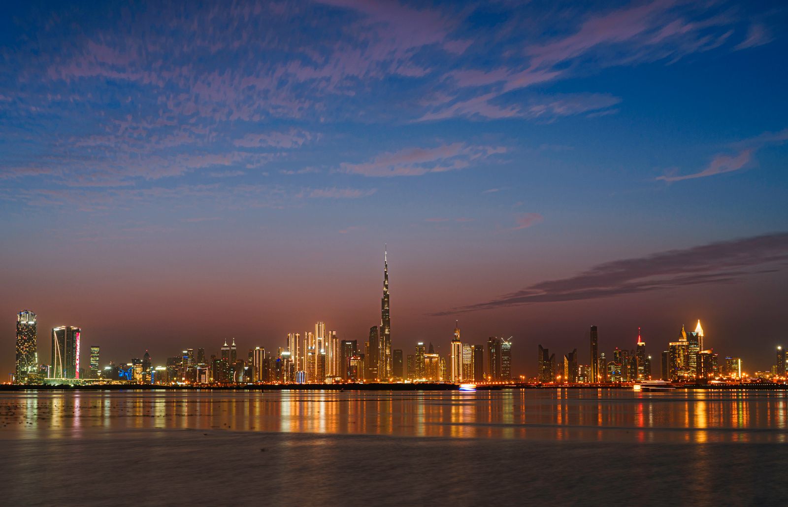 Downtown Dubai skyline at twilight, the illuminated Burj Khalifa spire standing tall above the city and reflected over the water