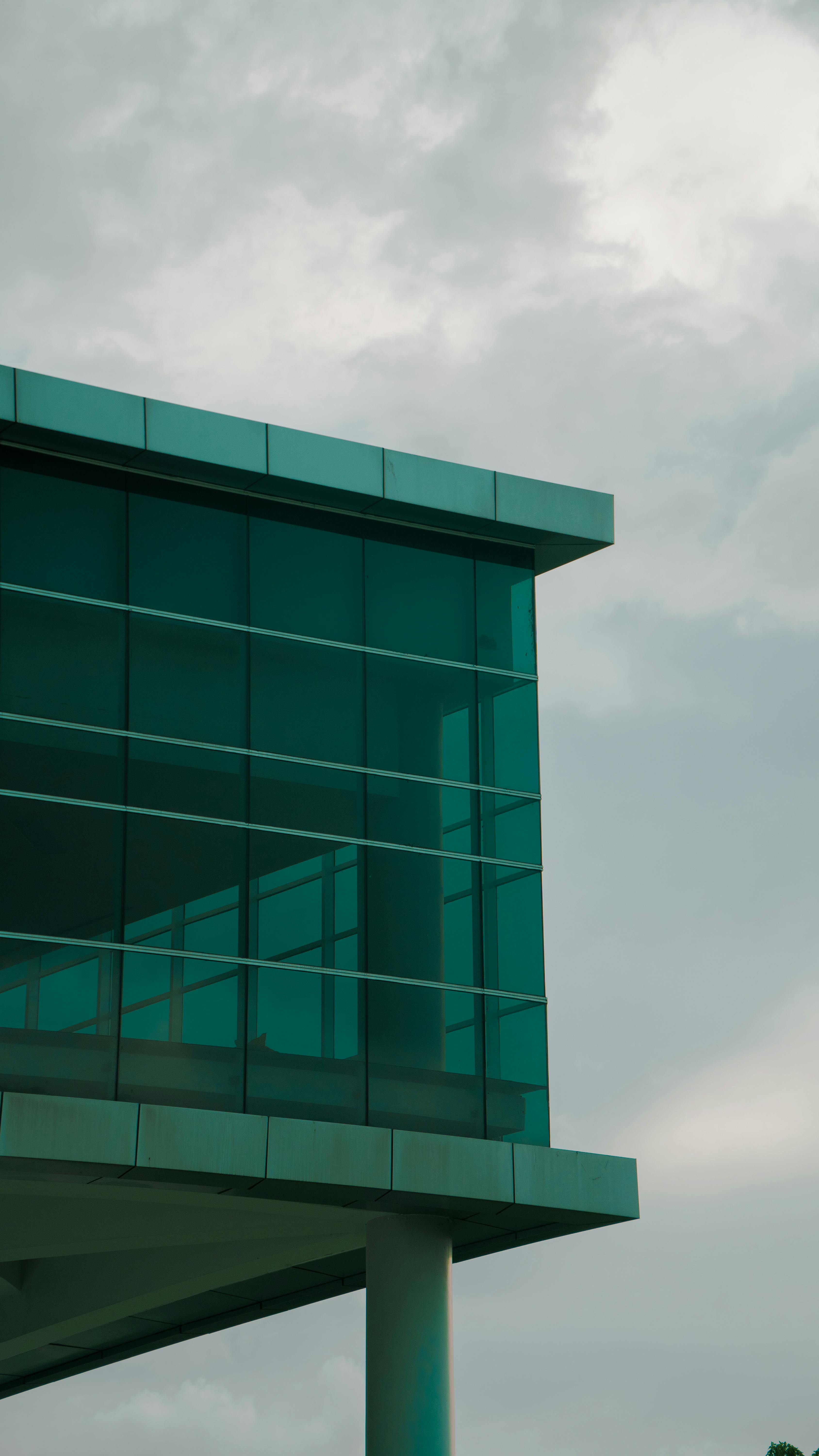 Free A corner view of a modern building featuring sleek glass panels against a cloudy sky. Stock Photo