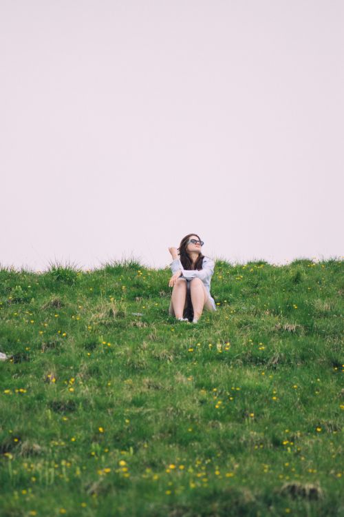 Free Woman in sunglasses sitting on a green hill under an overcast sky. Stock Photo