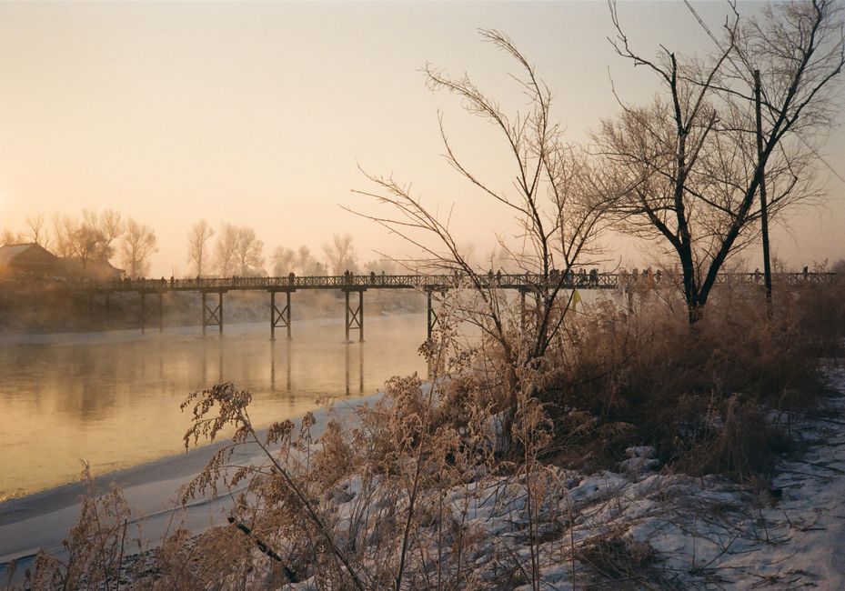 A tranquil winter scene with a footbridge over a river at dawn, showcasing frost-covered trees.