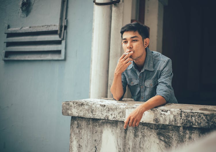 Thoughtful Young Asian Man Smoking Cigarette On Balcony