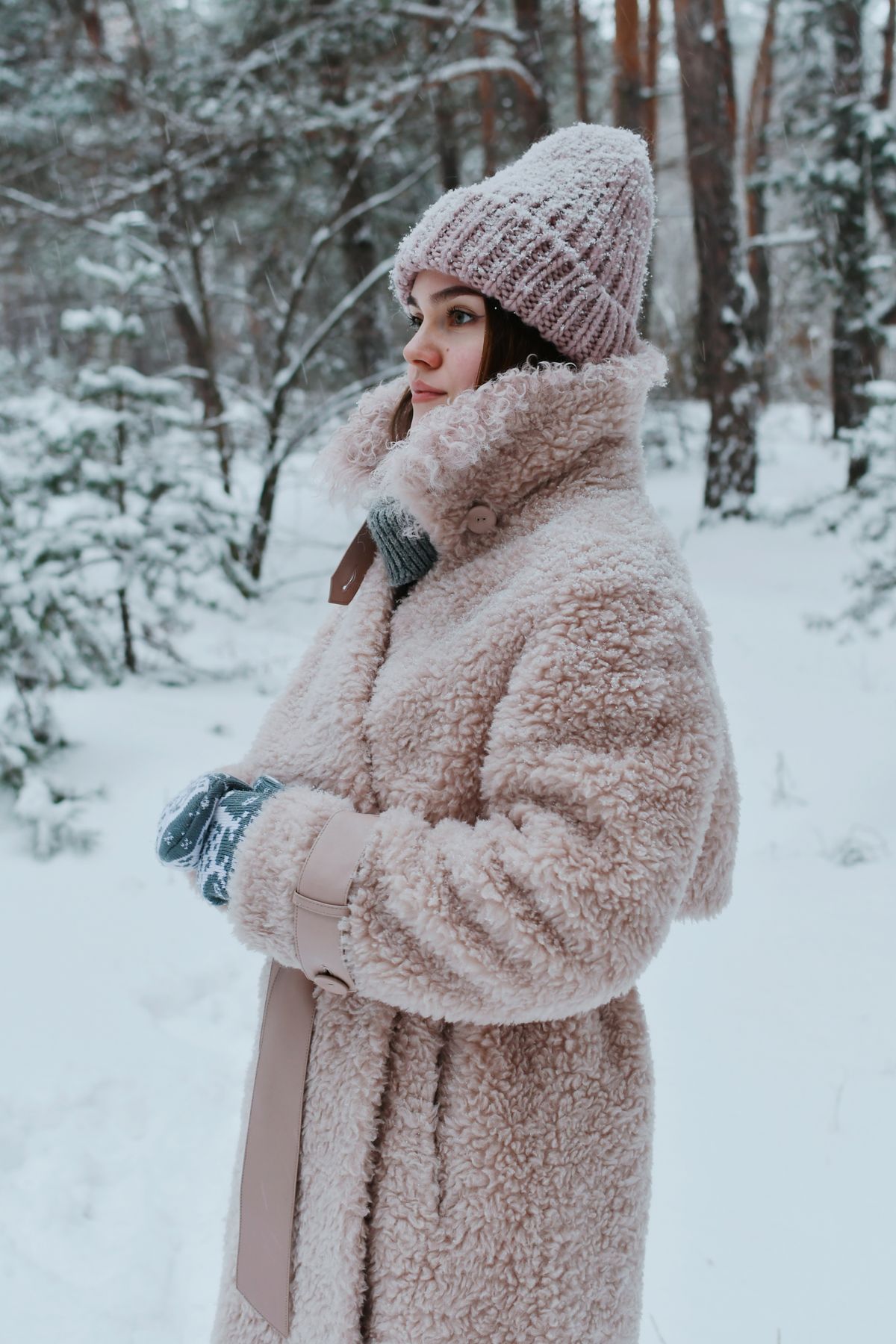 Woman in a belted blush winter coat with beanie standing in a snowy forest