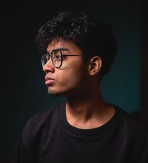 Free A thoughtful young man wearing glasses looks to the side in a dimly lit setting. Stock Photo