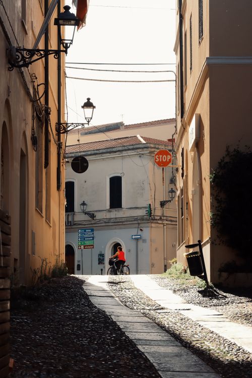 Free Explore the narrow cobblestone streets of Alghero, Sardinia, Italy, as a cyclist rides through with vintage architecture. Stock Photo