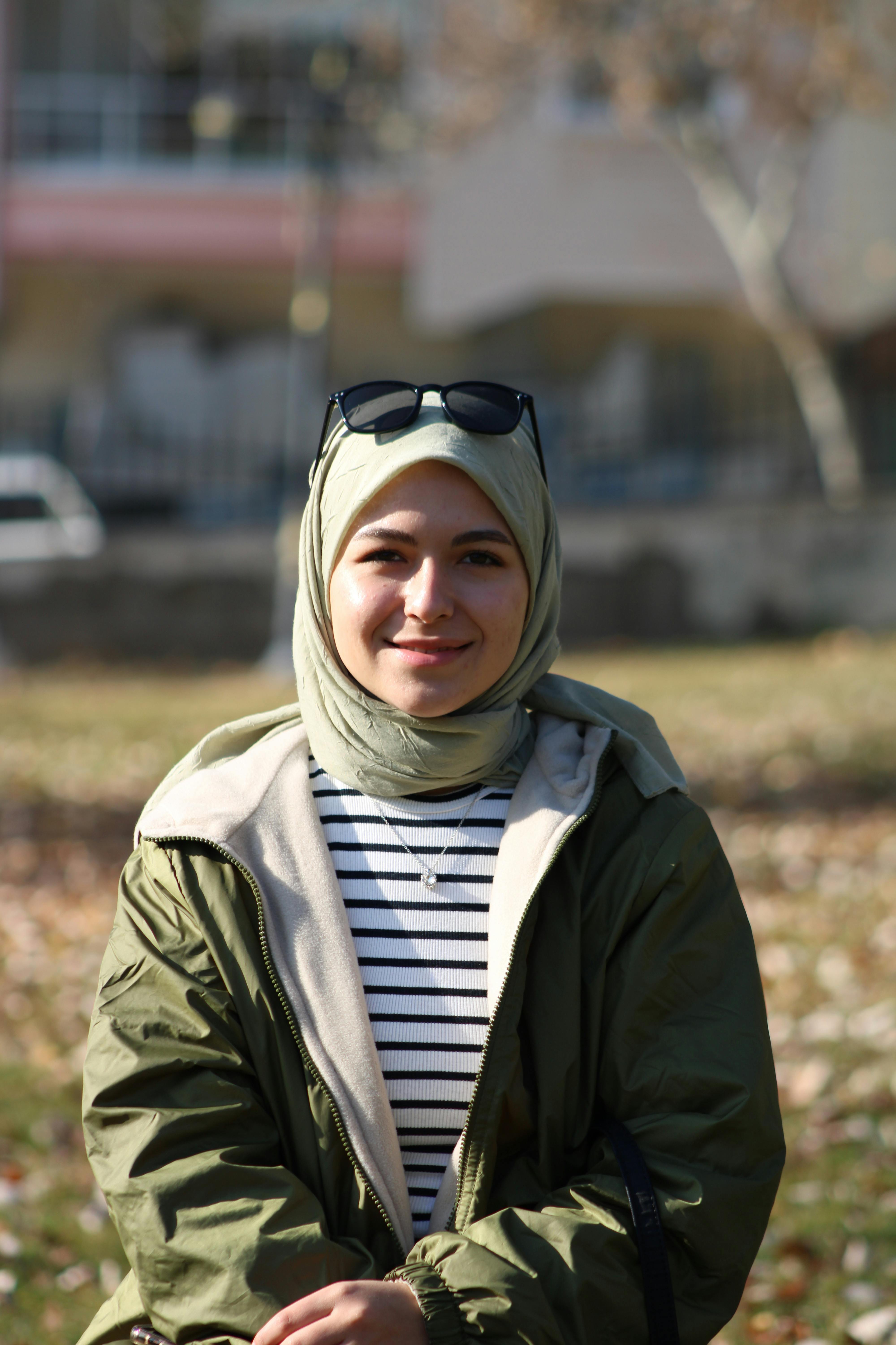 A smiling woman wearing a hijab and sunglasses, sitting outdoors on a sunny day.