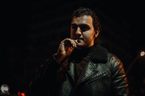Free A dramatic night portrait of a man in a black leather jacket smoking in an urban setting. Stock Photo