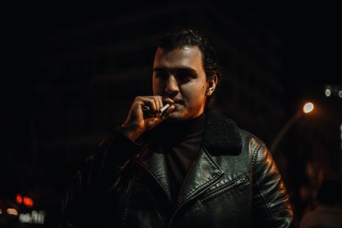 Free A man in a black leather jacket smoking a cigarette under streetlights at night. Stock Photo