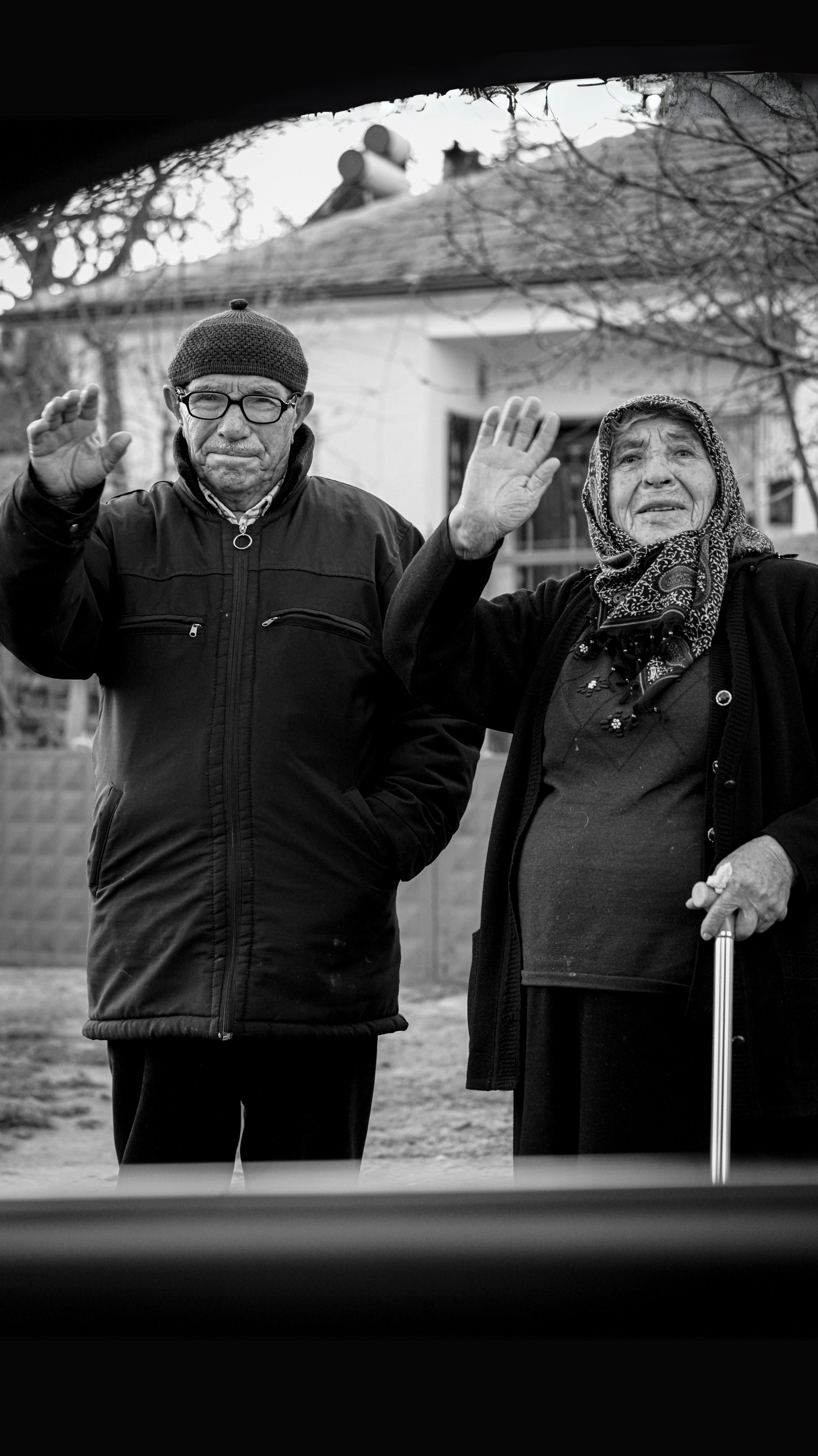 Black and white photo of an elderly couple waving, outdoors in Şereflikoçhisar, Ankara.