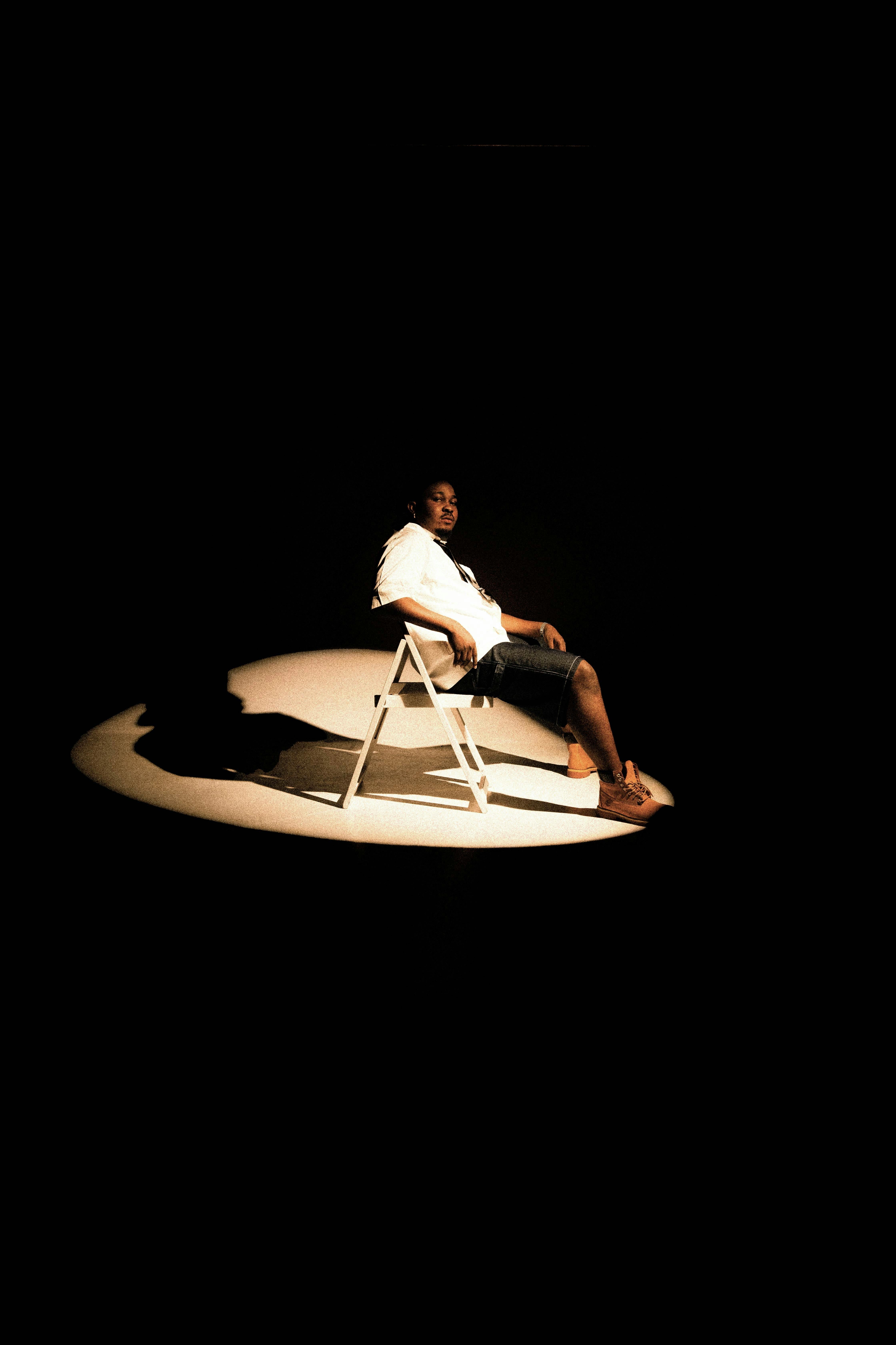 A man in a white shirt and shorts sits under a spotlight, creating a dramatic scene.