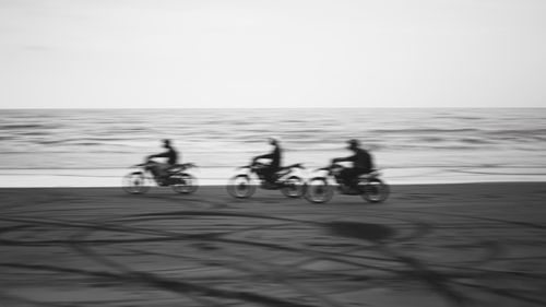 Free Black and white photo capturing bikers racing along a beach with blurred motion highlighting speed. Stock Photo
