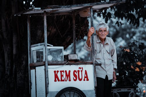 Elderly Man Standing next to a Food Cart on a Street 