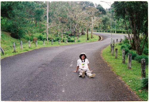 Free Young girl in casual attire sits relaxed on a rural road surrounded by lush greenery. Stock Photo