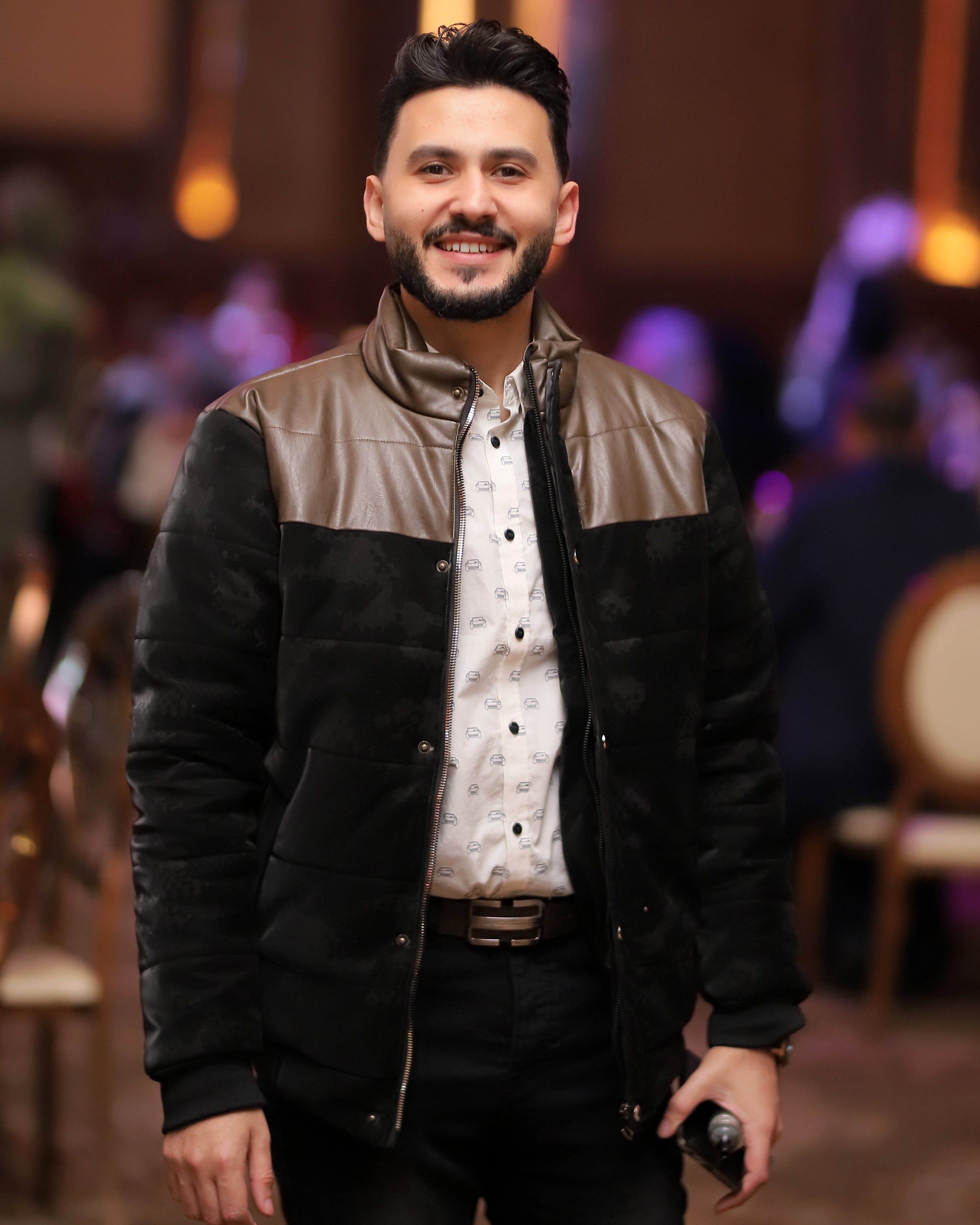 A stylish man wearing a fashionable jacket smiles warmly indoors, holding a microphone.