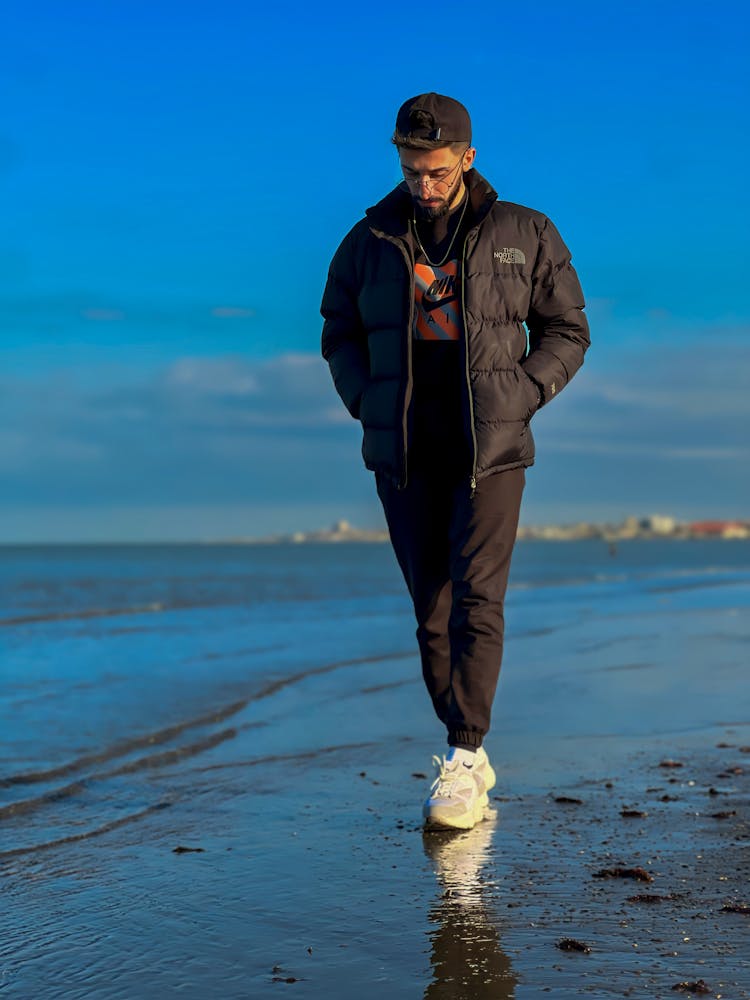 Bearded Man In Black Puffer Jacket Walking Along Beach