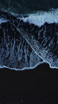A dramatic aerial shot capturing ocean waves crashing onto a dark sandy beach, showcasing the beauty of nature.