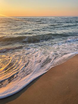Calm ocean waves wash over sandy shore at sunrise, creating a peaceful scene.