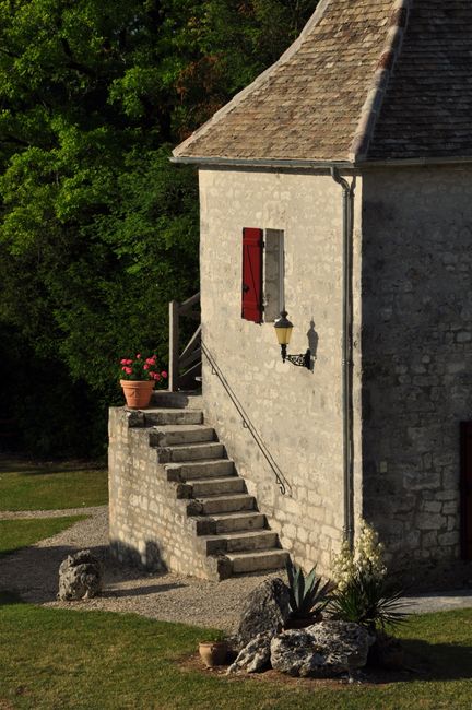 A quaint stone cottage with sunlit stairs and a red window shutter, surrounded by lush greenery.