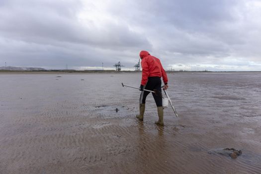 Man in red jacket and boots combs the beach with fishing gear, cloudy sky overhead.